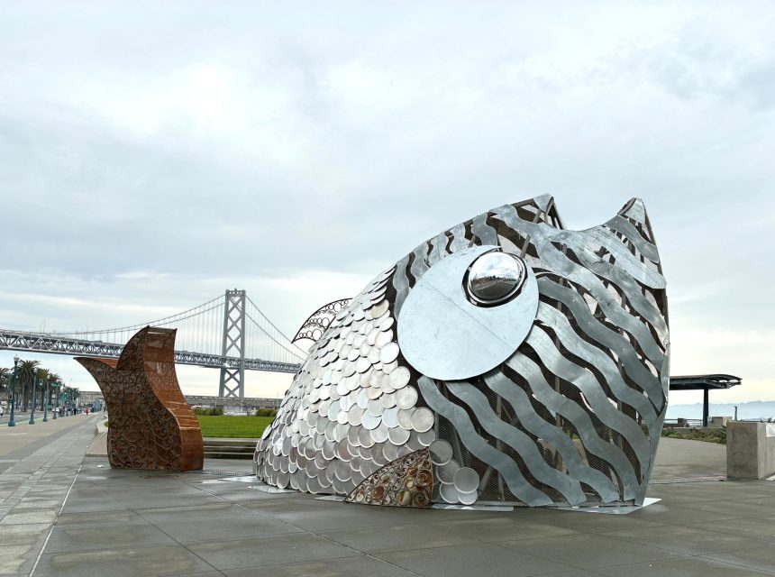 Large metallic fish sculpture with scales and wavy patterns stands on a waterfront promenade, with a bridge and cloudy sky in the background.