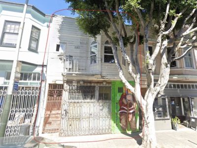 A two-story building with a metal gate, peeling white facade, green door, and a mural of a person in a brown coat, partially obscured by a tree on the sidewalk.