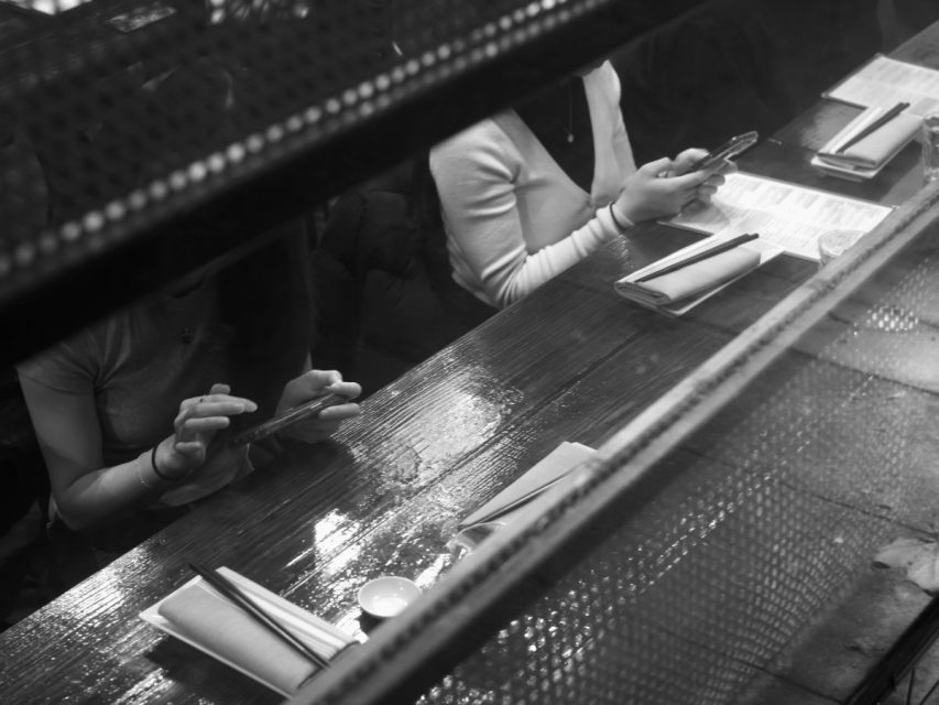 Two people sit at a wooden counter, each looking at their phone, with menus, chopsticks, and napkins placed in front of them.