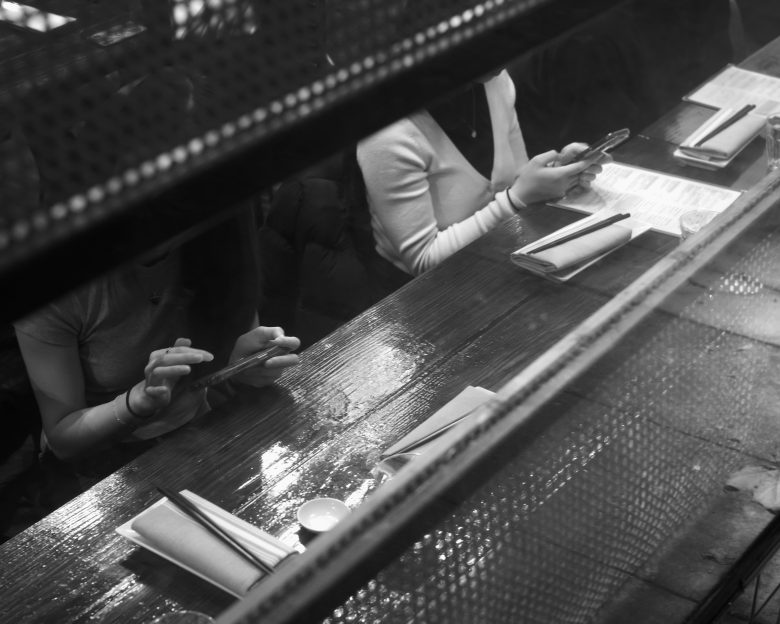 Two people sit at a wooden counter with chopsticks and menus, using their phones. The image is in black and white and viewed from above and behind a divider.