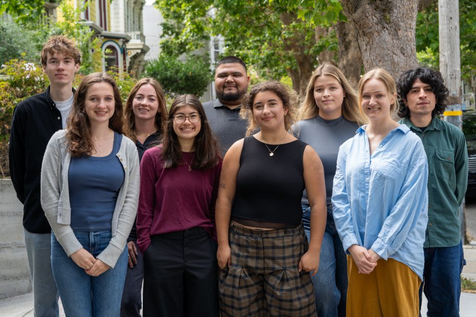 A group of nine people stands outdoors on a sidewalk in front of trees and houses, posing for a photo and facing the camera.