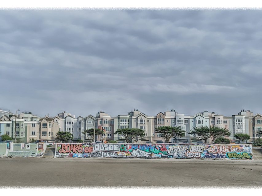 Row of pastel-colored houses behind a low wall covered in colorful graffiti, under a cloudy sky, with an empty sandy foreground.