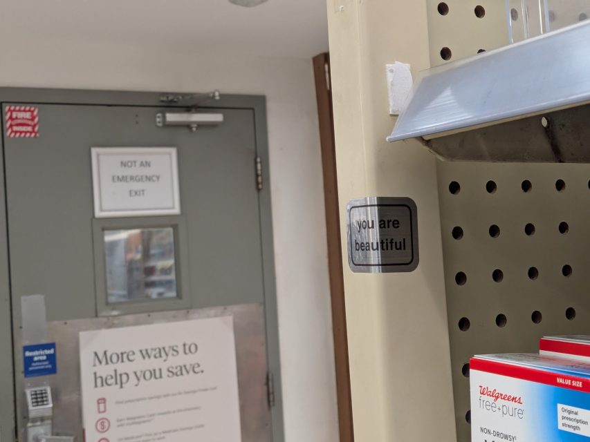 A store aisle shelf with a sign that reads "You are beautiful;" in the background, a door marked "Not an emergency exit" and a notice about savings.