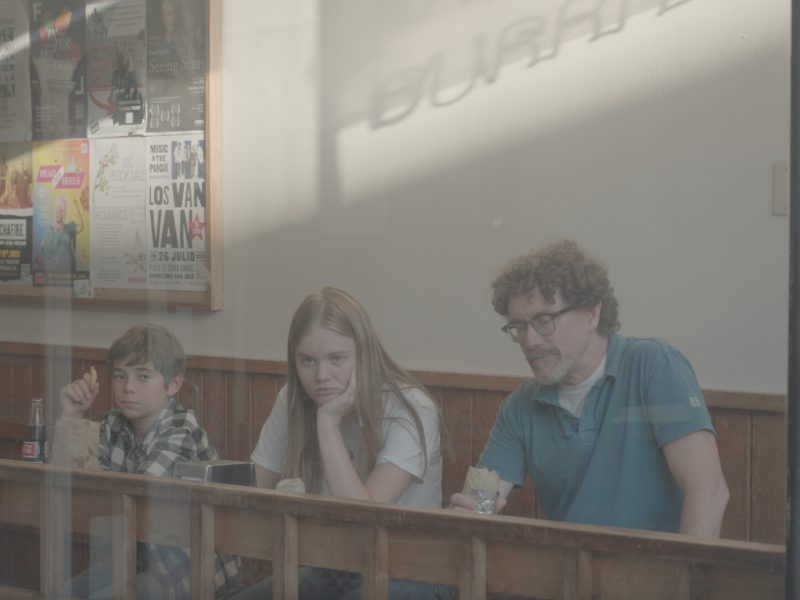 Three people sit at a counter inside a restaurant, each holding food, with neutral or bored expressions. Posters decorate the wall behind them.