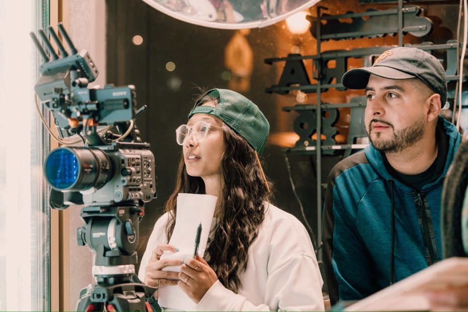 Two people stand behind a professional video camera; one holds a notepad and pen, both appear focused on the filming process.