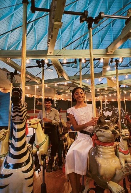 Two people riding carousel animals indoors; one is on a zebra and the other on a cat, with string lights and a blue ceiling visible above.