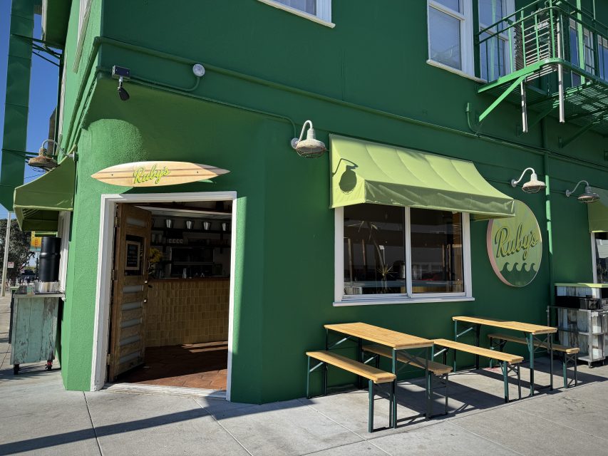 A green building with a yellow-green awning and sign reading "Ruby's," outdoor wooden tables, and an open entrance to a café.