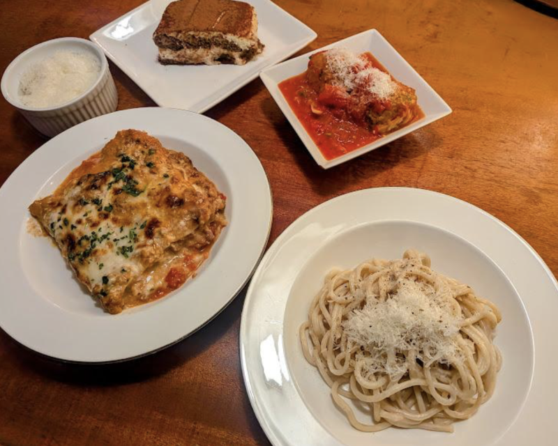 Plates of lasagna, spaghetti with grated cheese, stuffed cannelloni in tomato sauce, a bowl of grated cheese, and a slice of tiramisu on a wooden table.
