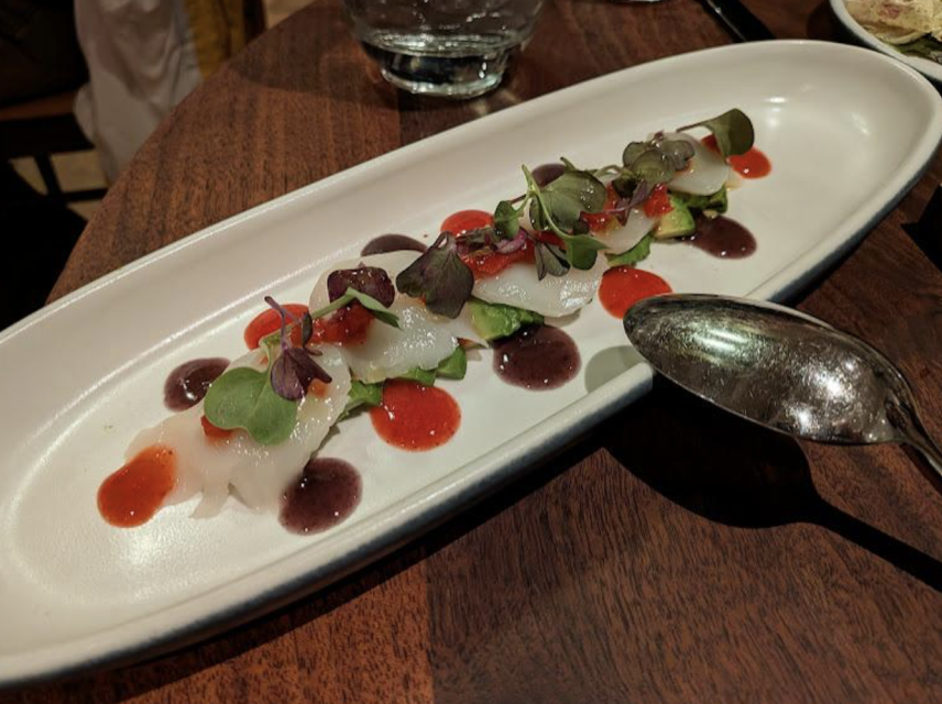 Thin slices of raw fish topped with microgreens and red and purple sauces are arranged on a long white plate next to a metal spoon.