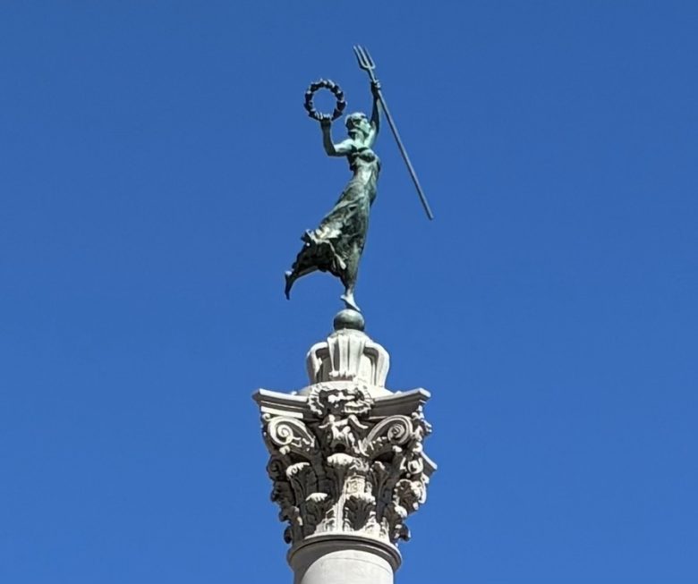 Bronze statue of a woman holding a wreath and a trident stands atop an ornate stone column against a clear blue sky.