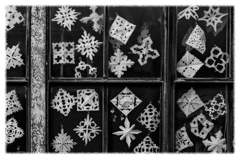 Black-and-white photo of window panes covered with various paper snowflakes and geometric cut-out shapes taped to the glass.