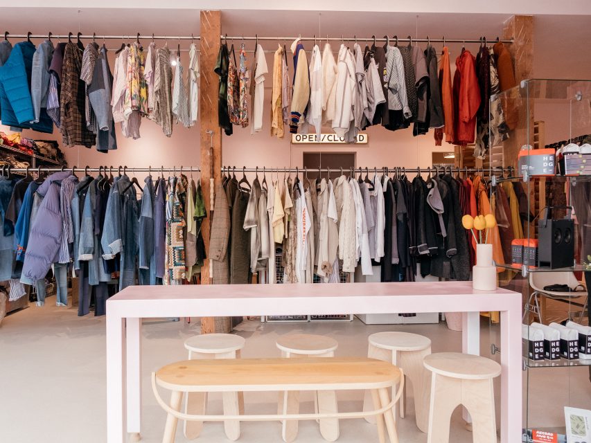 Clothing store interior with racks of assorted jackets and shirts, a long pink table with stools, and neatly arranged merchandise on shelves.
