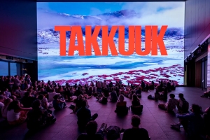 A large group of people sits on the floor in front of a big screen displaying a snowy landscape and the word "TAKKUUK" in bold red letters.