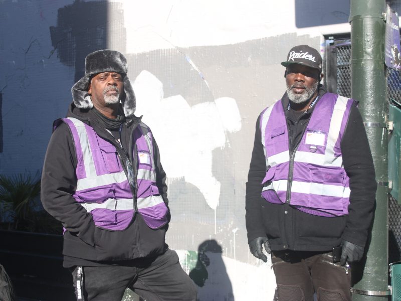 Two men wearing black clothing and purple reflective vests stand on a city street in sunlight, with a painted wall and green pole behind them.