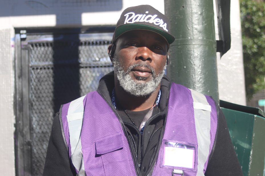 A man with a gray beard wearing a Raiders cap and a purple safety vest stands outdoors near a green pole and a fenced area.