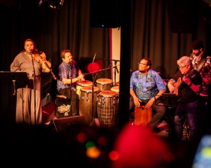 A band performs on stage with a female singer, two percussionists, a guitarist, and another musician, all illuminated by stage lights.