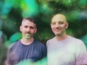 Two men stand side by side outdoors in front of greenery, both wearing casual t-shirts. The image appears slightly blurred with colorful light effects.