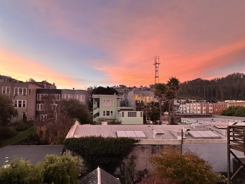 Sunset over a neighborhood with houses, apartment buildings, trees, and hills in the background under a colorful sky with pink and orange clouds.