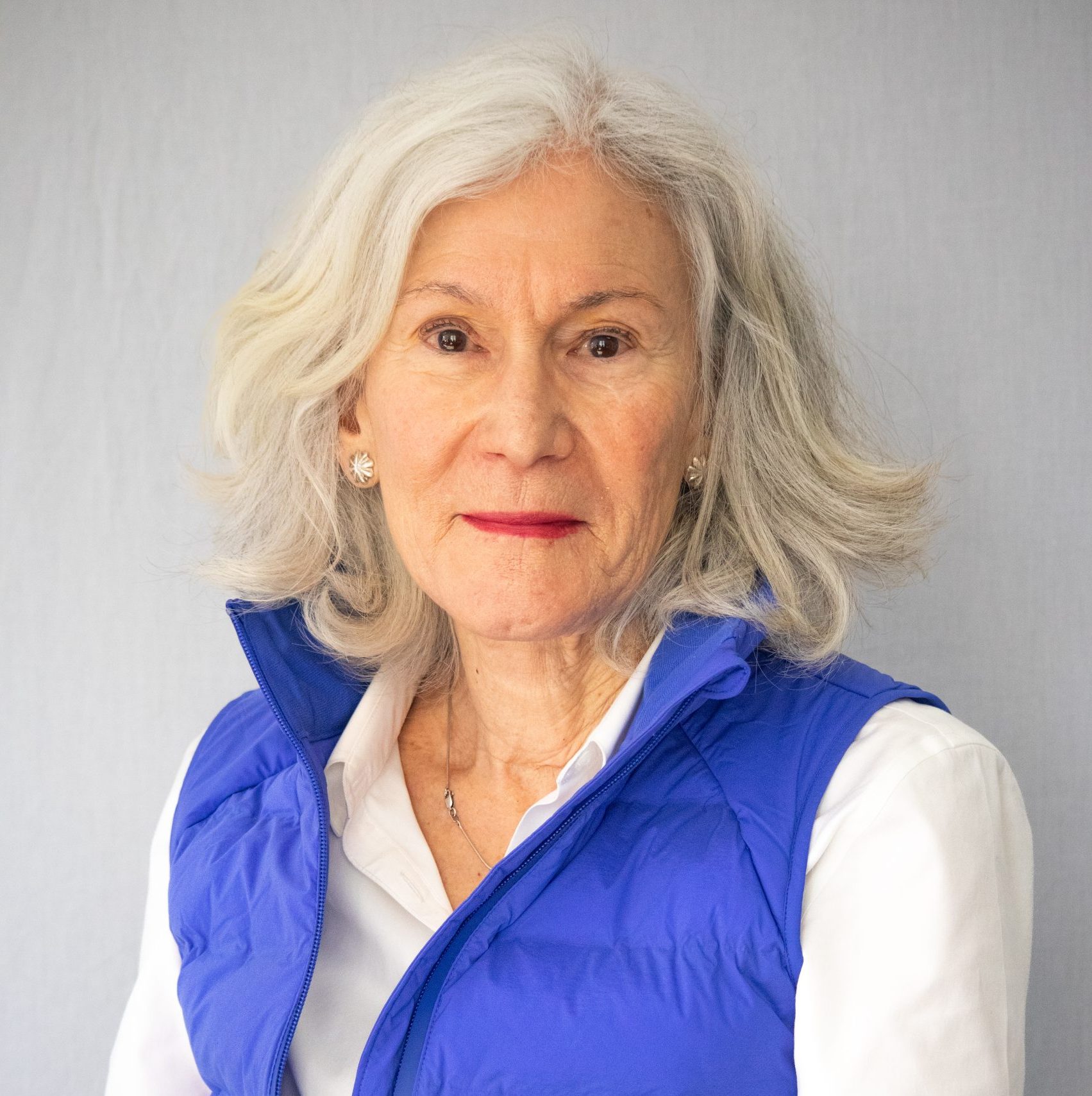 An older woman with grey hair, wearing a white shirt and blue vest, poses in front of a plain light background.