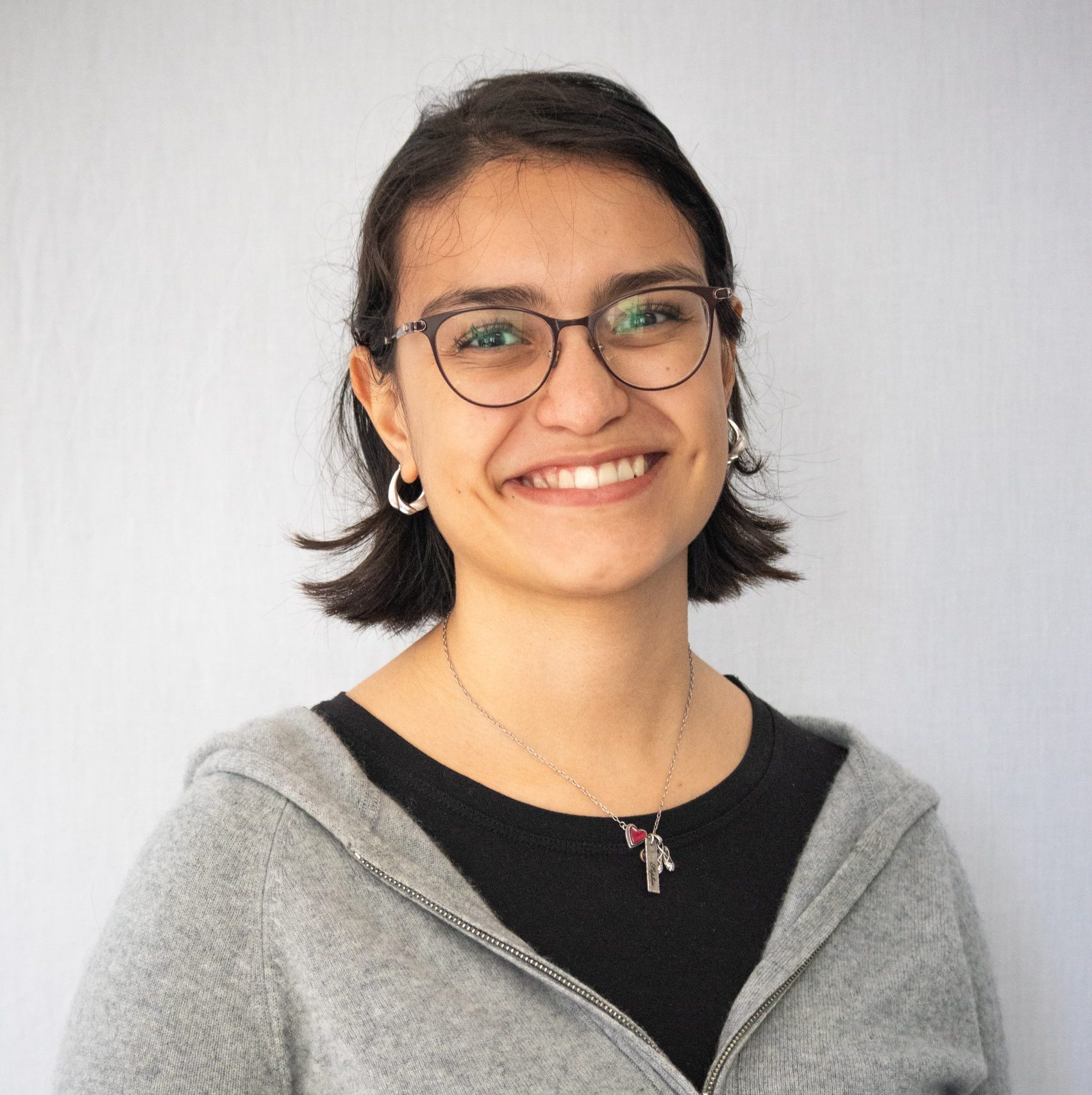 A person with short dark hair, glasses, and a grey hoodie smiles at the camera against a plain light background.