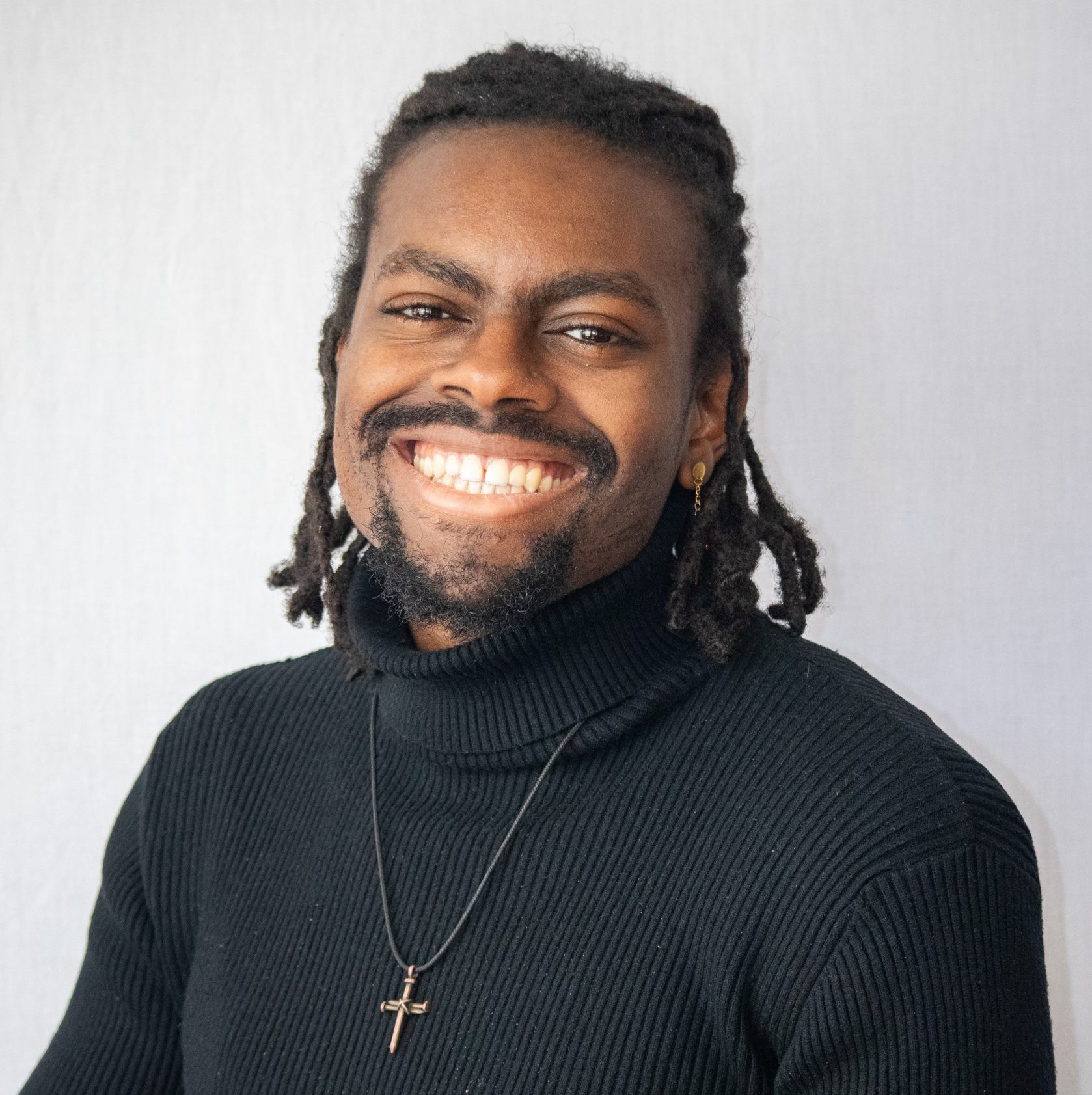 A person with medium brown skin, wearing a black turtleneck and a cross necklace, smiles at the camera against a light background.