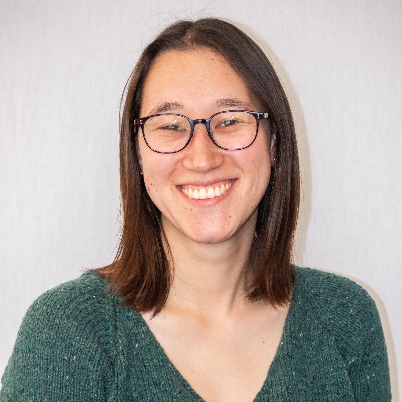 A woman with straight brown hair and glasses smiles at the camera, wearing a green sweater against a plain light-colored background.