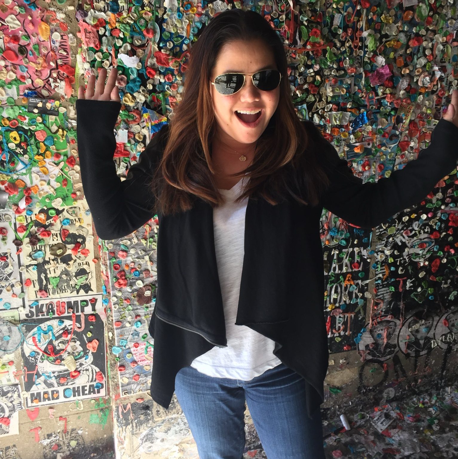 A person wearing sunglasses stands in front of a colorful wall covered in chewed gum, raising their hands and smiling.