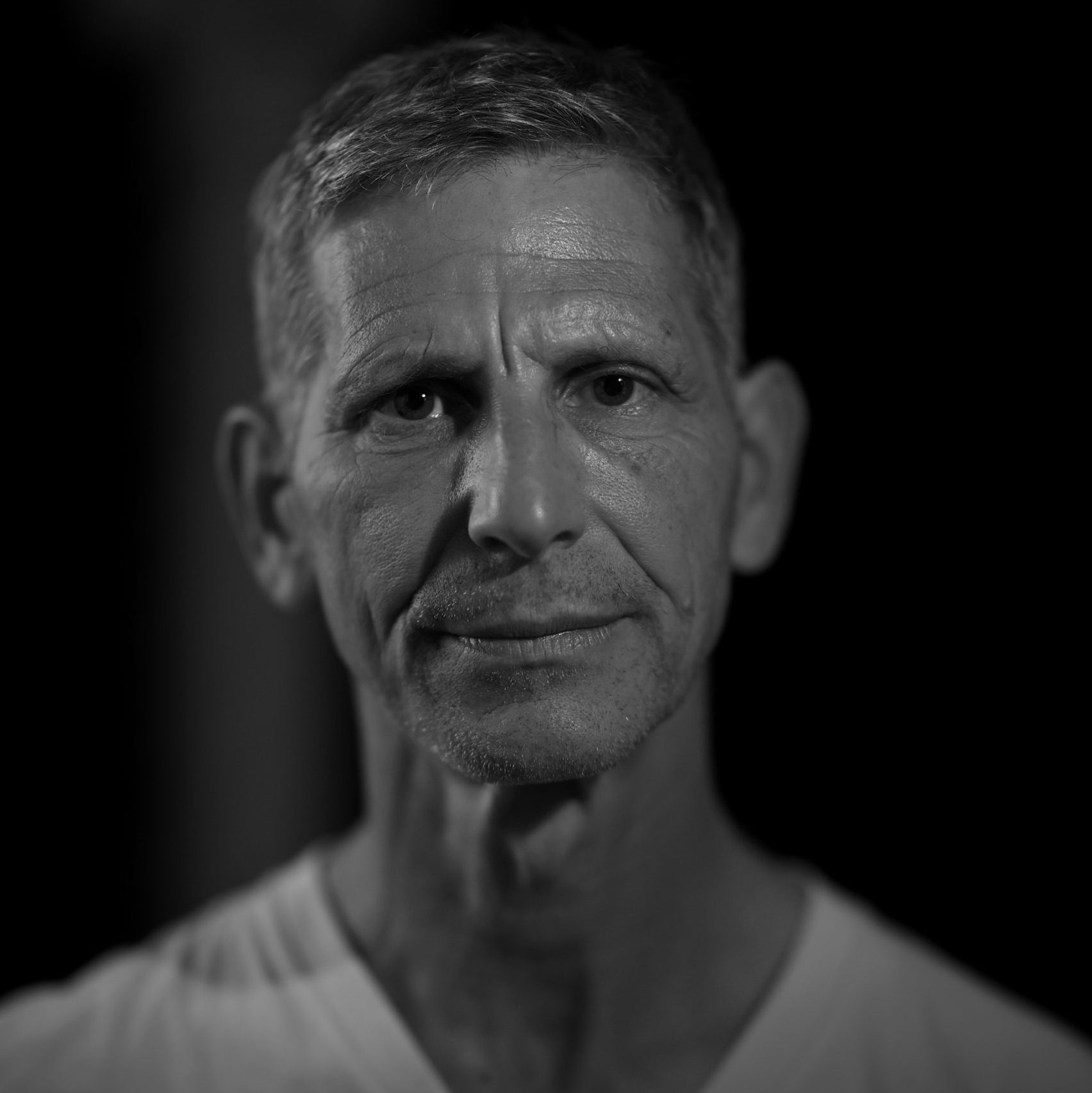 A middle-aged man with short gray hair and a serious expression, wearing a plain white shirt, is photographed in black and white against a dark background.