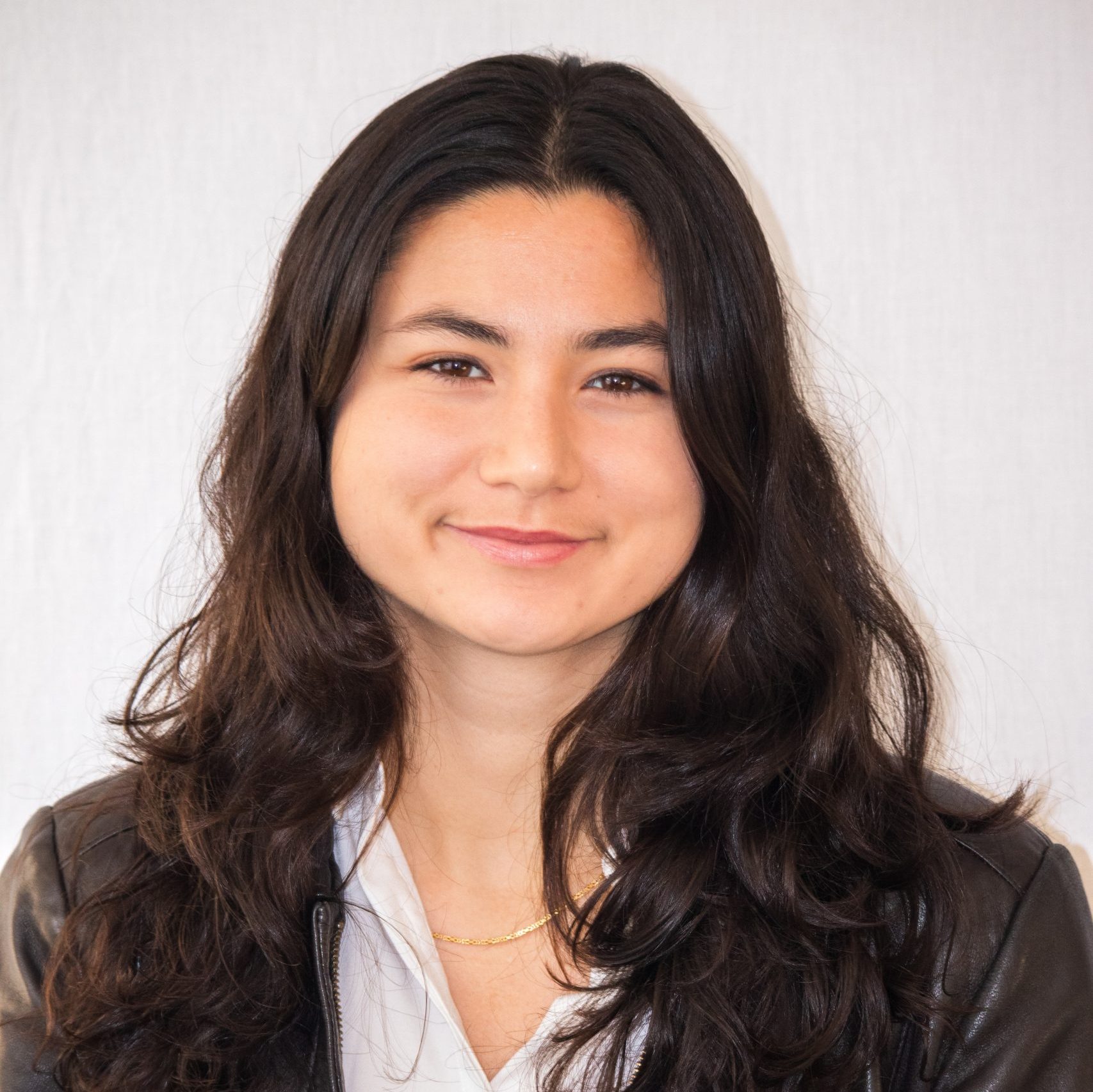A young woman with long dark hair, wearing a white shirt and brown jacket, smiles at the camera against a plain light background.