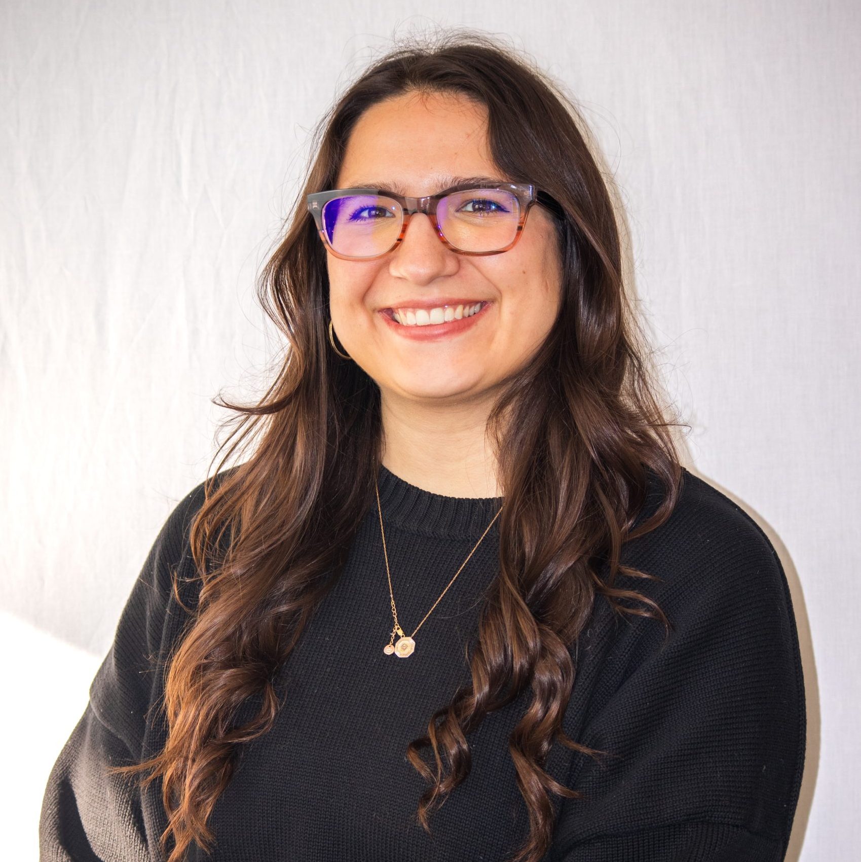 A woman with long brown hair and glasses, wearing a black sweater and layered necklaces, smiles in front of a plain light background.