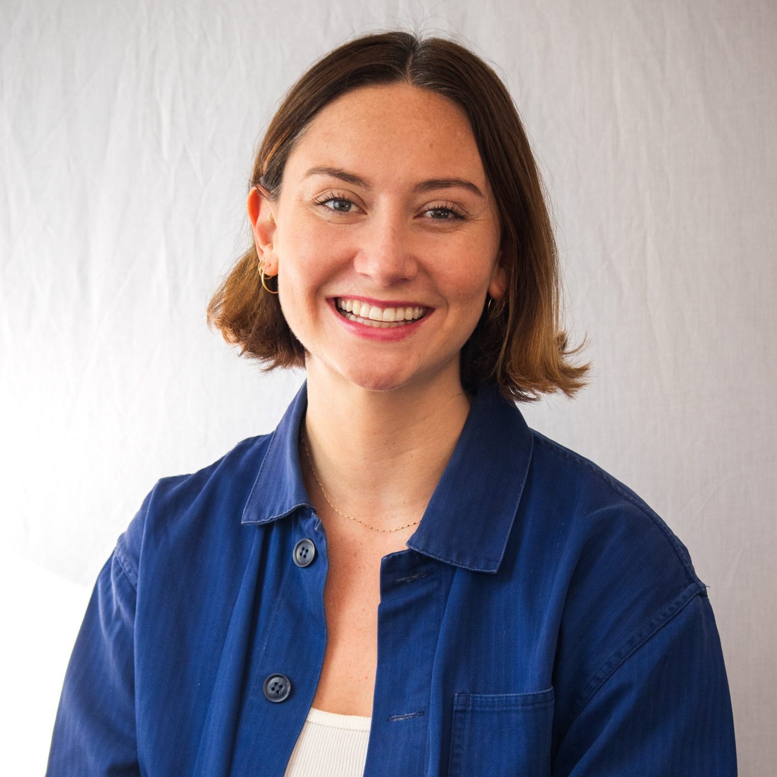 A woman with short brown hair, wearing a blue button-up shirt over a white top, smiles at the camera against a plain light background.