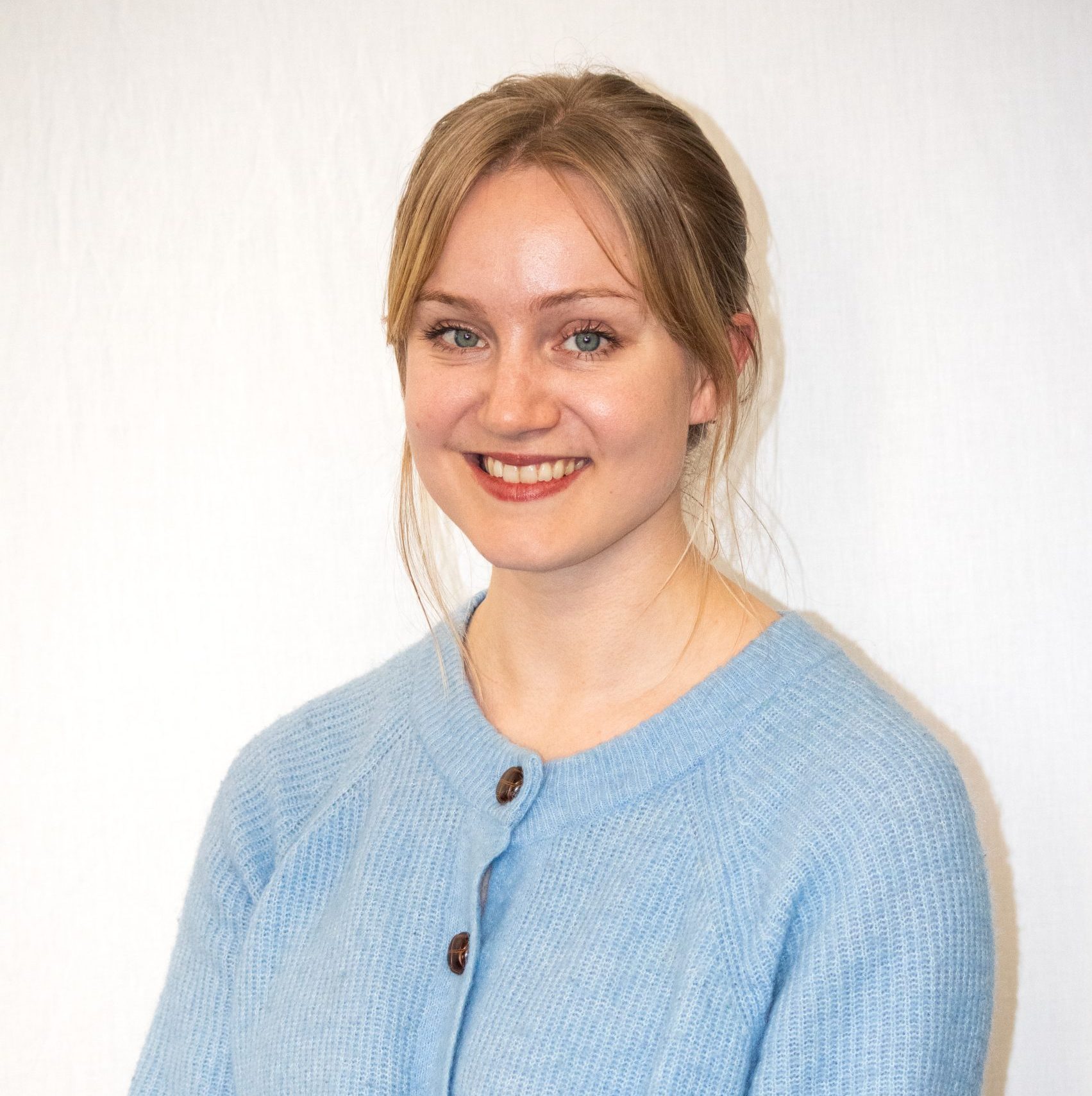 A woman with blonde hair tied back, wearing a light blue sweater, is smiling at the camera against a plain white background.