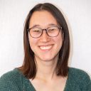 A person with straight brown hair, wearing glasses and a green sweater, smiles at the camera in front of a plain light background.