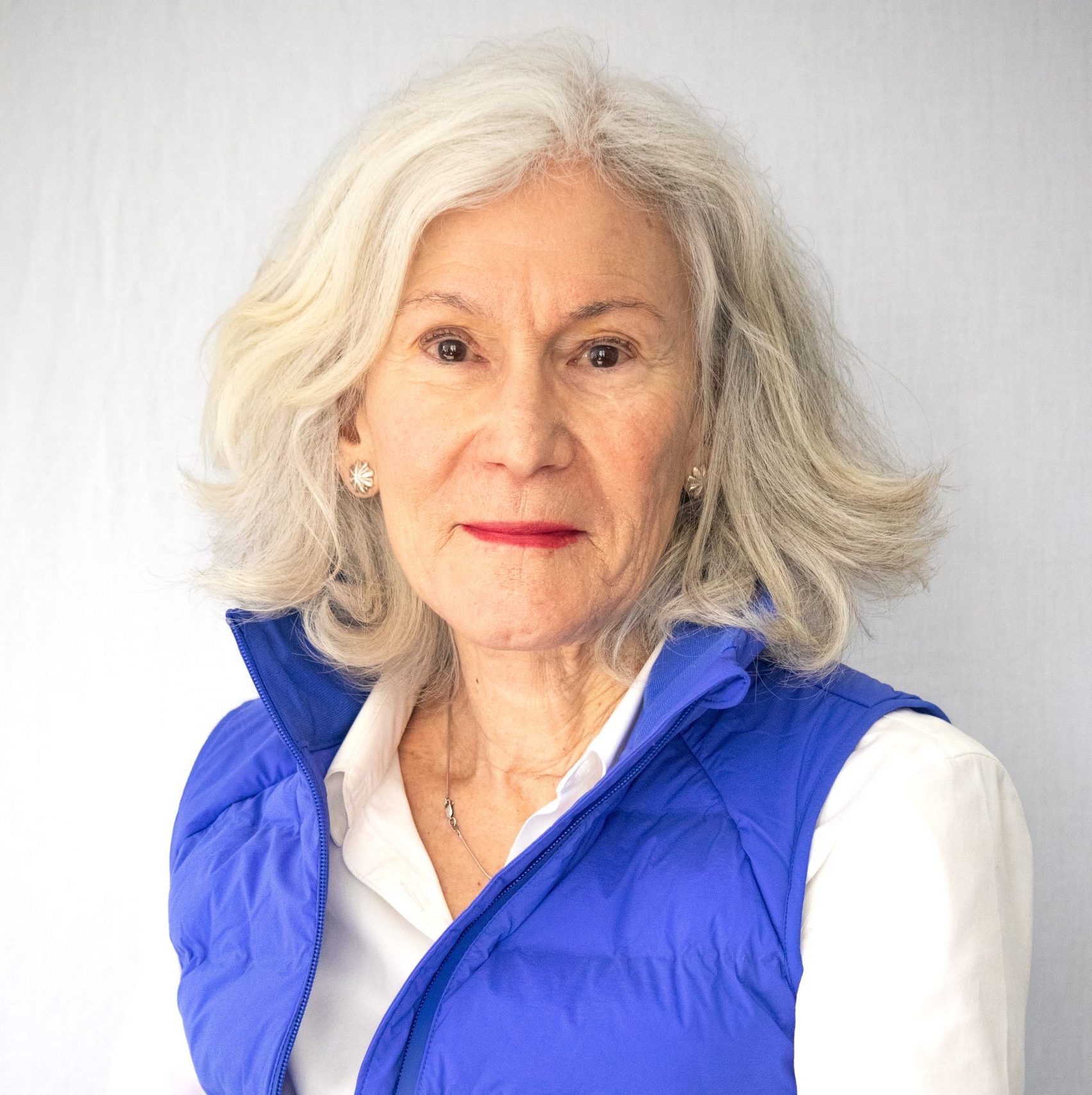 An older woman with gray hair wearing a blue vest over a white shirt, standing in front of a light background and looking at the camera.