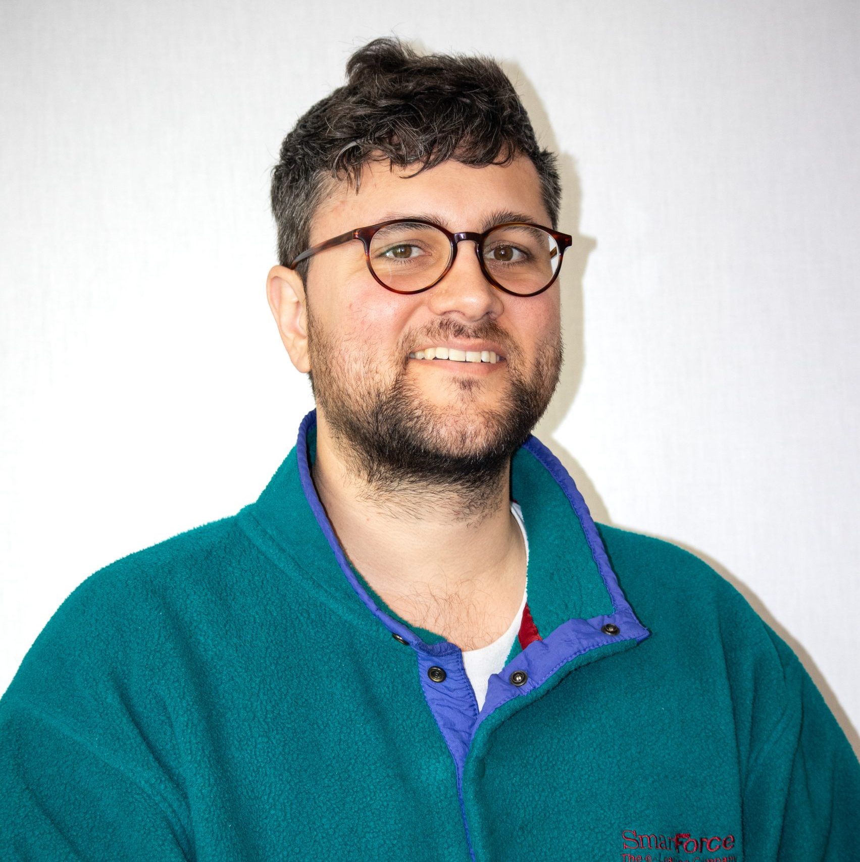 A person with short dark hair, a beard, and glasses, wearing a teal fleece jacket, smiles in front of a plain light background.