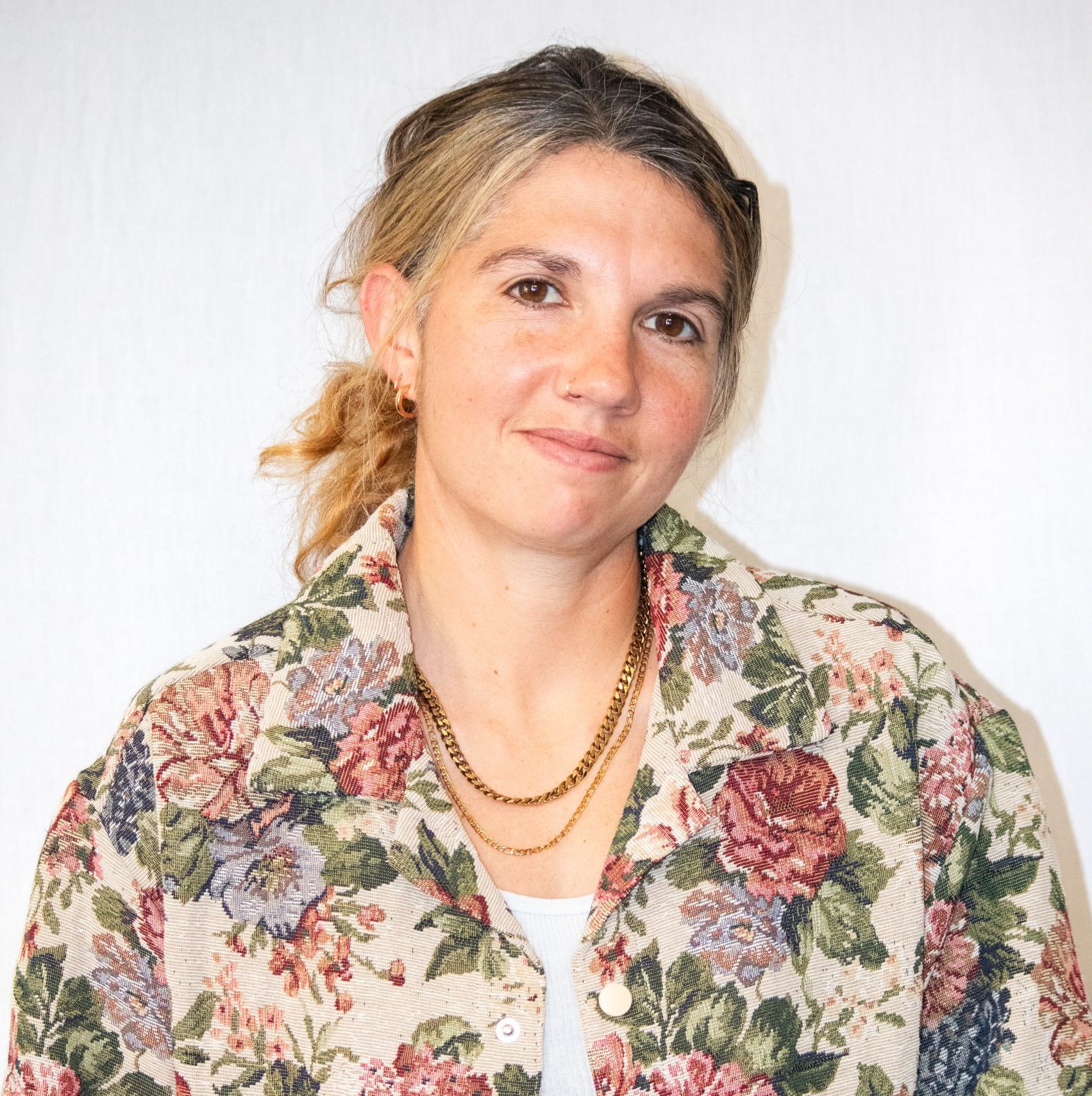 A woman with light brown hair in a ponytail wears a floral-patterned jacket, a white top, and gold necklaces, standing against a plain white background.