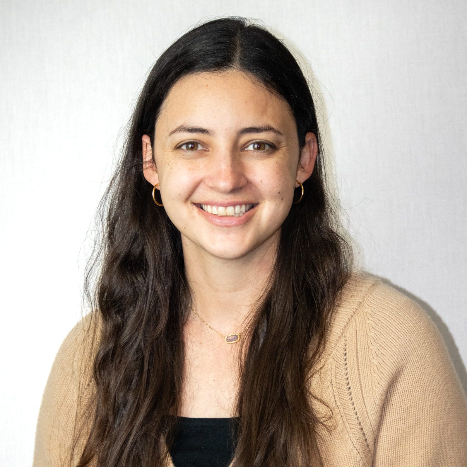 Woman with long dark hair, wearing a tan sweater and small hoop earrings, smiling in front of a plain light background.