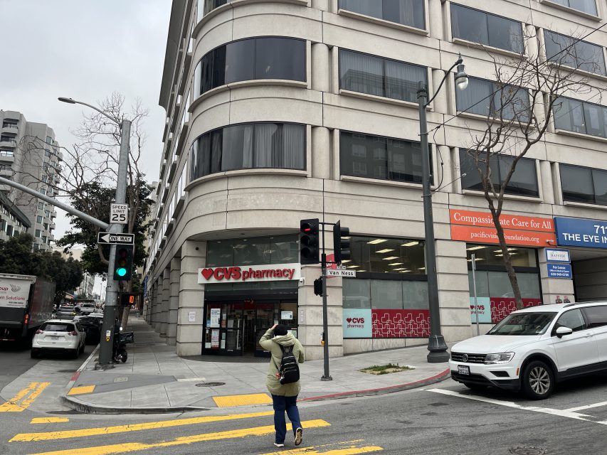 Person crossing the street toward a CVS Pharmacy location on the ground floor of a large building at a city intersection.