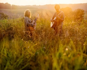 Two people stand in a sunlit field playing guitars, surrounded by tall grass and warm, golden light.