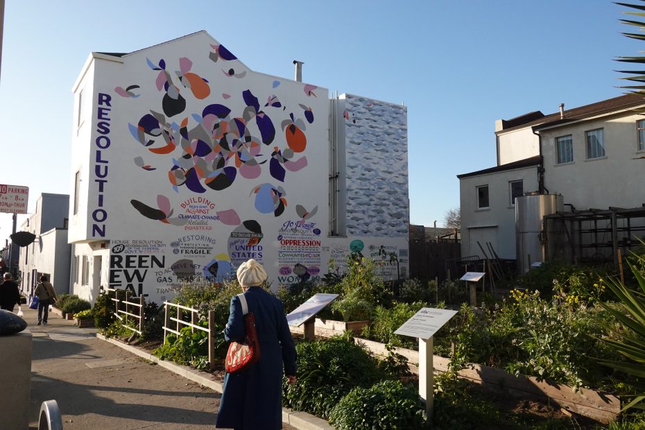 A person walks by a community garden with signs in front of a white building featuring colorful abstract murals and the word "RESOLUTION" painted vertically.
