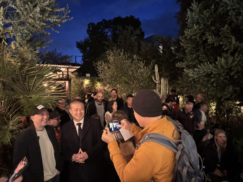 A person in an orange jacket takes a photo of a smiling man in a suit amid a group of people outdoors at night, surrounded by trees and lights.