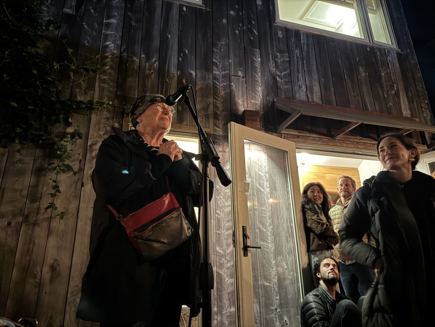A woman speaks into a microphone outside a wooden building at night, while several people watch from the doorway and nearby.