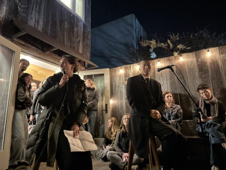 Two people sit and speak into microphones at an outdoor night event, with an audience seated and standing around them under string lights.