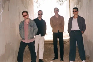 Four men wearing sunglasses stand in a sunlit tunnel, posing casually with hands in pockets and neutral expressions.