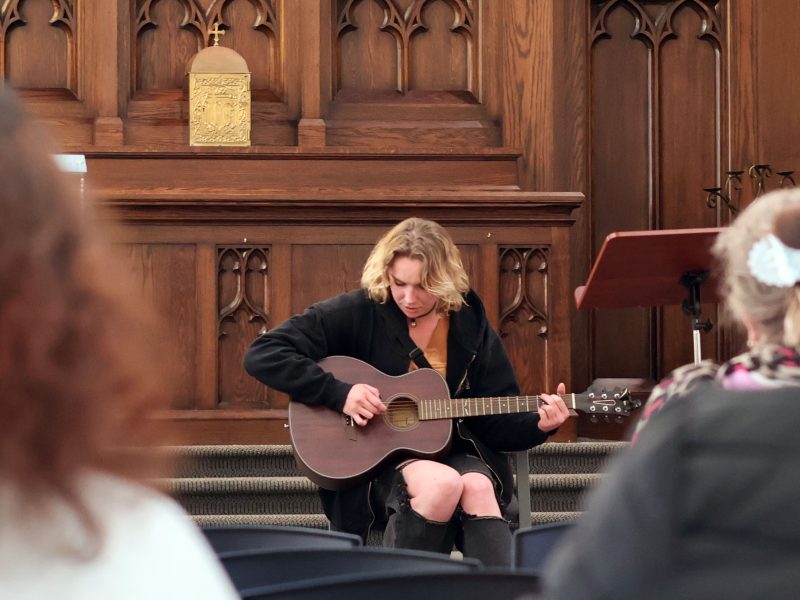 A person with shoulder-length blond hair plays an acoustic guitar while seated on a stage in a wooden-paneled room, facing an audience.