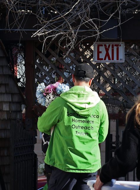 A person in a green "San Francisco Homeless Outreach Team" jacket holds a bouquet of flowers, standing near an "EXIT" sign and wooden lattice.