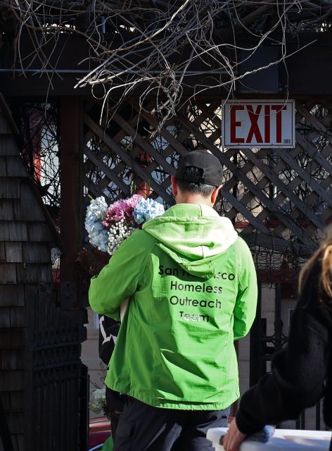 A person in a green “San Francisco Homeless Outreach Team” jacket holds a bouquet of flowers, standing near a gate under an “EXIT” sign.