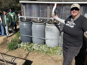 A person stands smiling beside three large rain barrels connected to a gutter system, with a group of people and a garden in the background.