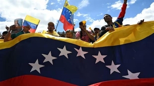 A group of people holds a large Venezuelan flag and smaller flags, standing outdoors under a partly cloudy sky.