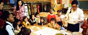 A teacher demonstrates an activity to a group of young children seated and standing around a table in a classroom.